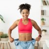 Successful Weight Loss. Happy African Woman After Slimming Showing Her Old Oversize Pants Standing Indoors, Smiling To Camera